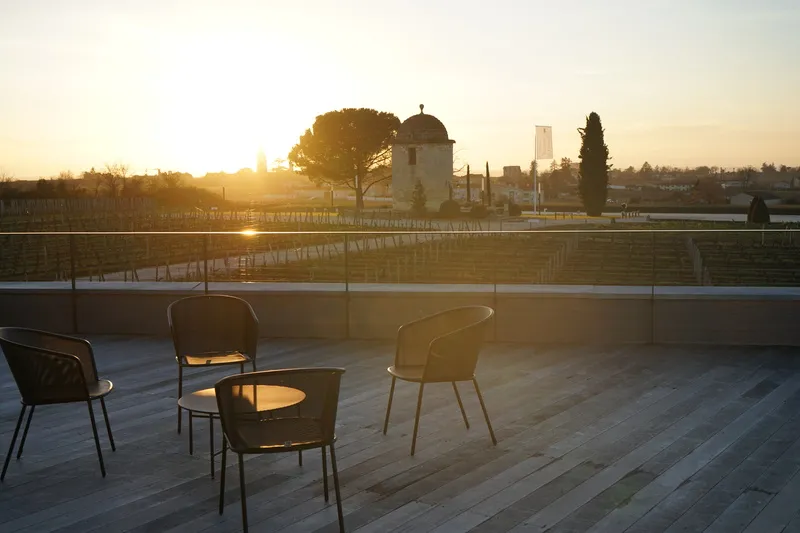 Modern seating area on a wooden terrace overlooking Bordeaux vineyards at sunset, with Château Balestard La Tonnelle in the background