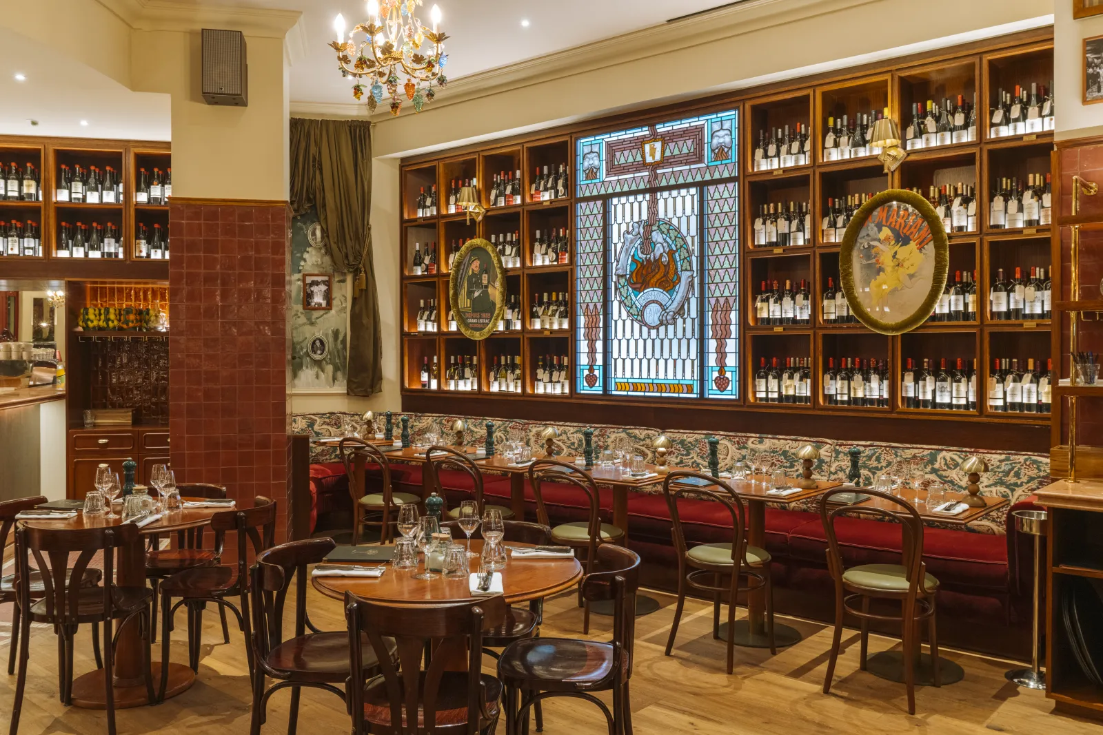 Elegant interior of Brasserie Vinatier in Bordeaux featuring wooden shelves stocked with wine bottles, round wooden tables, red velvet chairs, and classical décor with stained glass and gold ornaments.