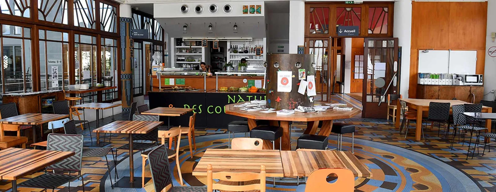 Modern interior space of Bordeaux's Les Bains with wooden furniture, low tables, black chairs, orange and black service counter, large windows, geometric colored flooring, and welcome signage.