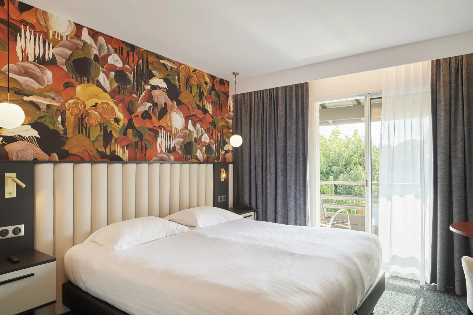 Modern hotel room featuring a queen-size bed with white linens, beige tufted headboard, vibrant orange floral wallpaper above the bed, gray curtains, glass doors opening to a balcony overlooking greenery, and contemporary lighting fixtures.