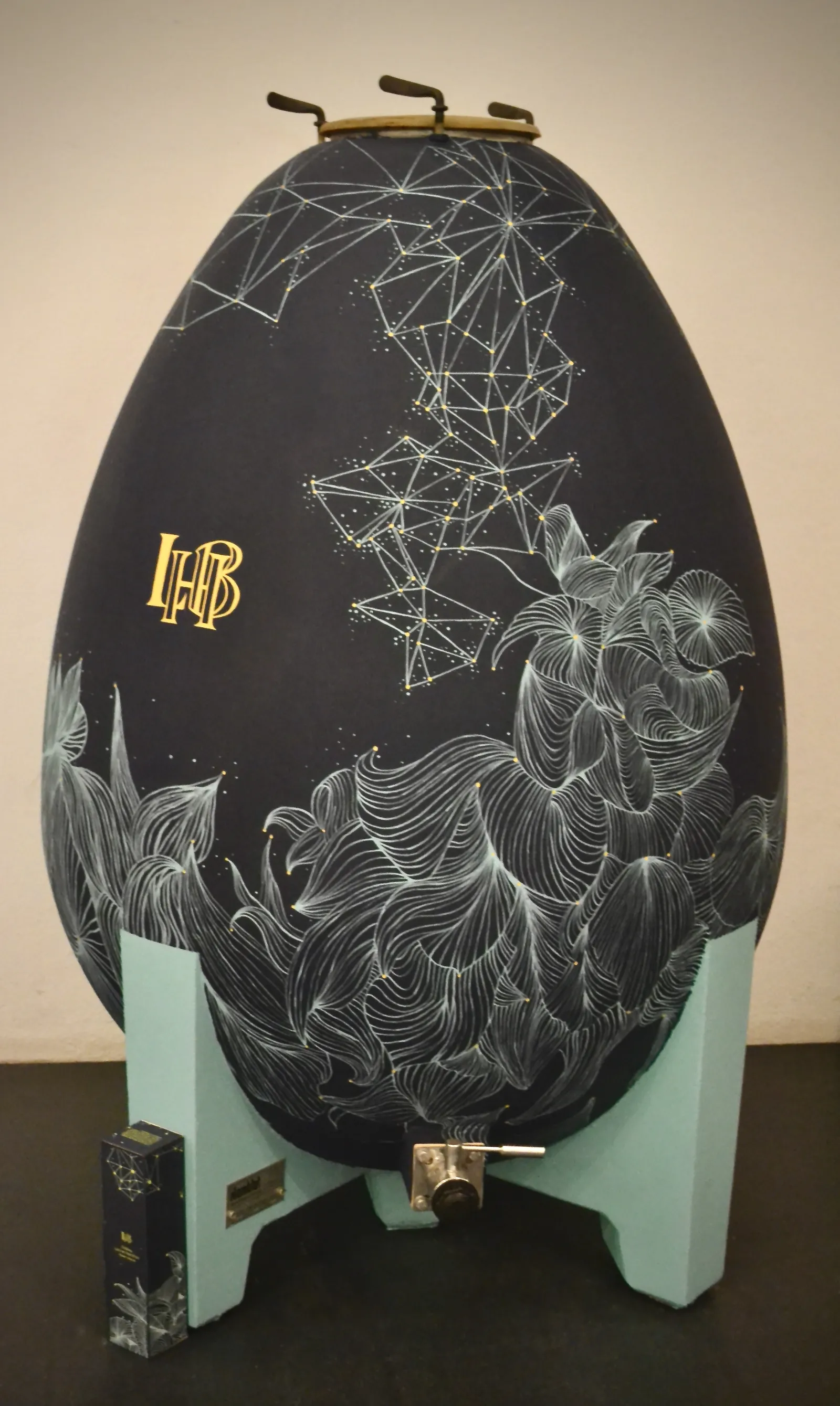 Black decorative Easter egg from Château Larrivet Haut-Brion in Bordeaux, featuring engraved flowers and gold geometric patterns, displayed on a turquoise stand.