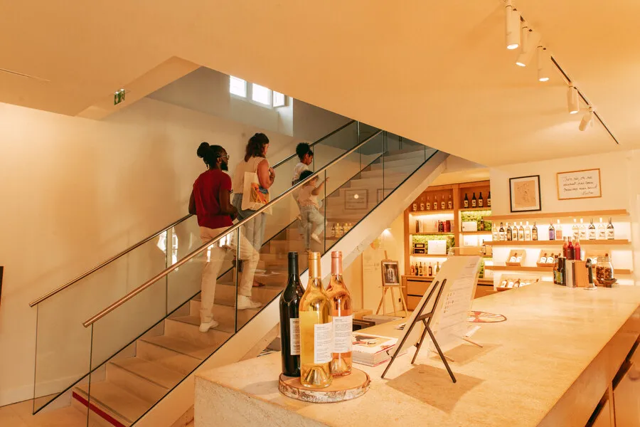 Modern interior of Château Toulouse-Lautrec in Bordeaux showing three visitors climbing a glass staircase, with rosé wine bottles displayed in the foreground and illuminated shelves of wine products in the background.