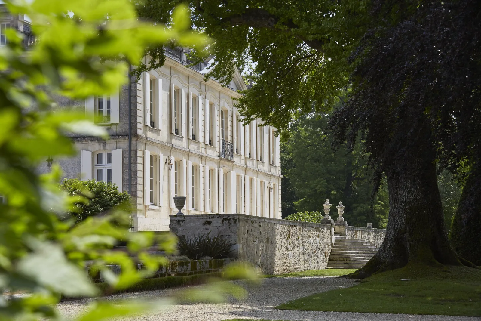 View of Château de La Dauphine in Bordeaux featuring its white neoclassical facade with columns, surrounded by lush greenery and ancient trees, accessed by stone steps.