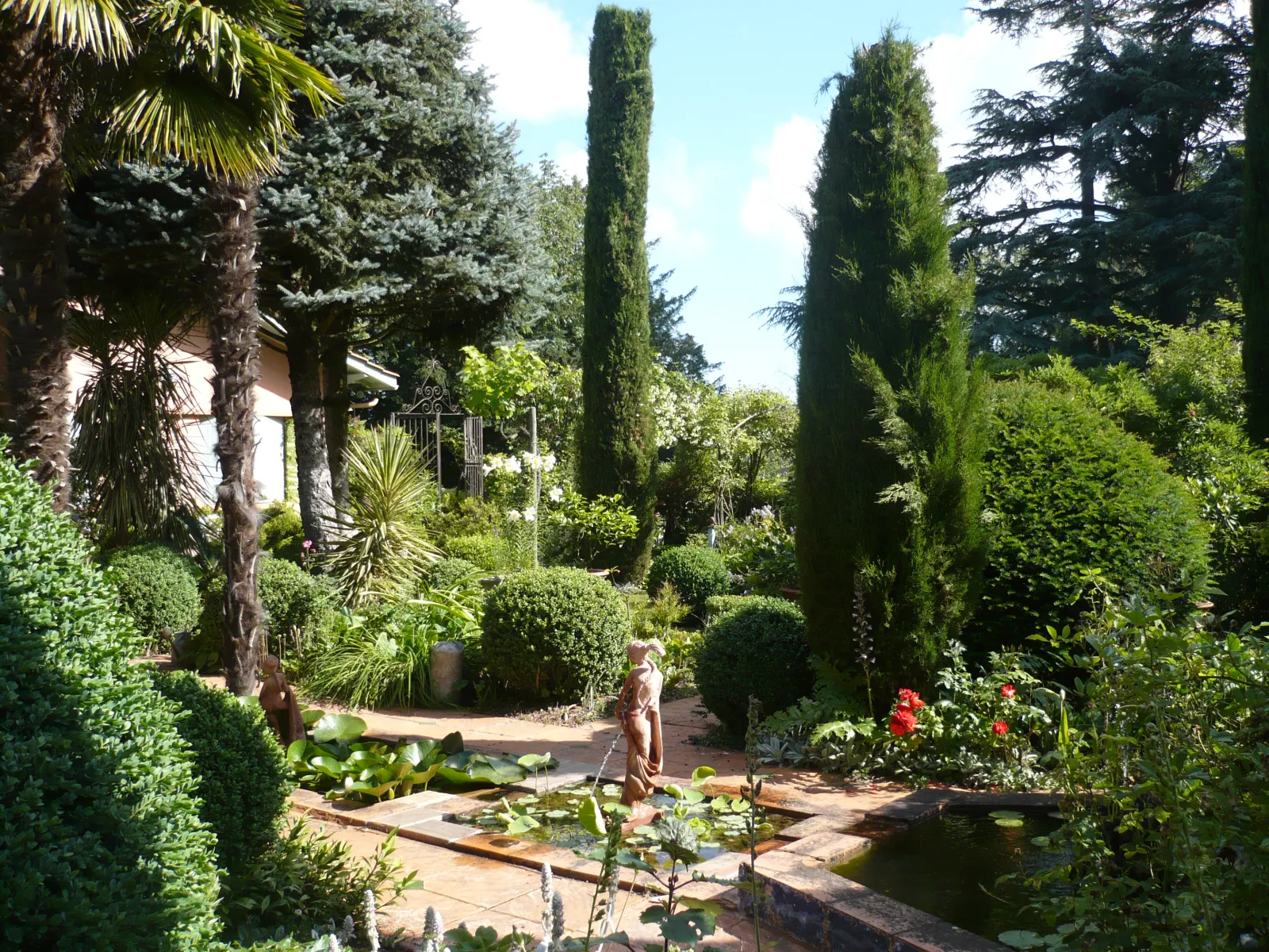 The Clozet Garden in Bordeaux, a landscaped garden featuring brick pathways, a water basin, cypress trees, palms and sculptures surrounded by lush greenery.