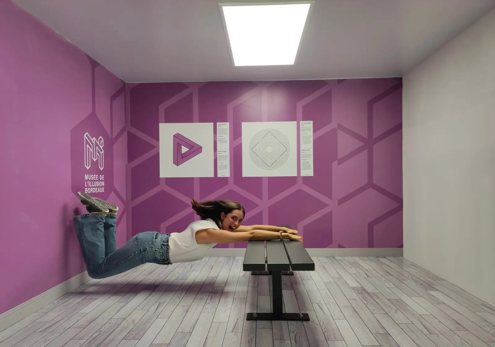A woman lying on the floor of a pink room at the Bordeaux Museum of Illusion, posing next to a black bench and interacting with an optical illusion, with geometric artworks displayed on the wall.