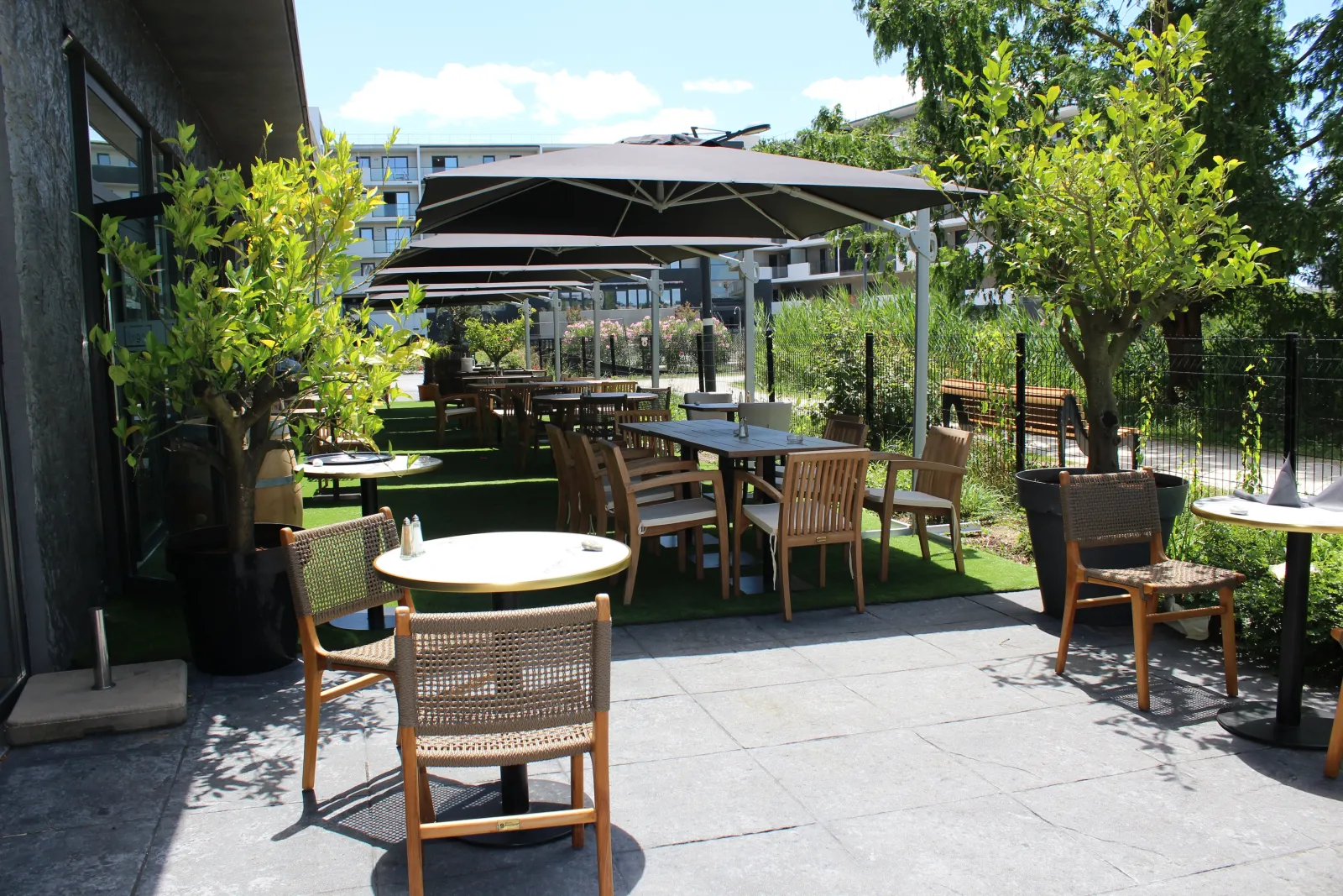 Outdoor terrace with round tables, wooden and woven chairs, black umbrellas, artificial grass, greenery and modern buildings in the background in Bordeaux.