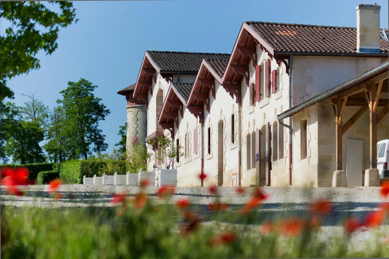 The Maison des Hôtesses in Bordeaux, a historic white stone building with red-tiled roofs, red shuttered windows and a side tower, surrounded by red flowers in the foreground.