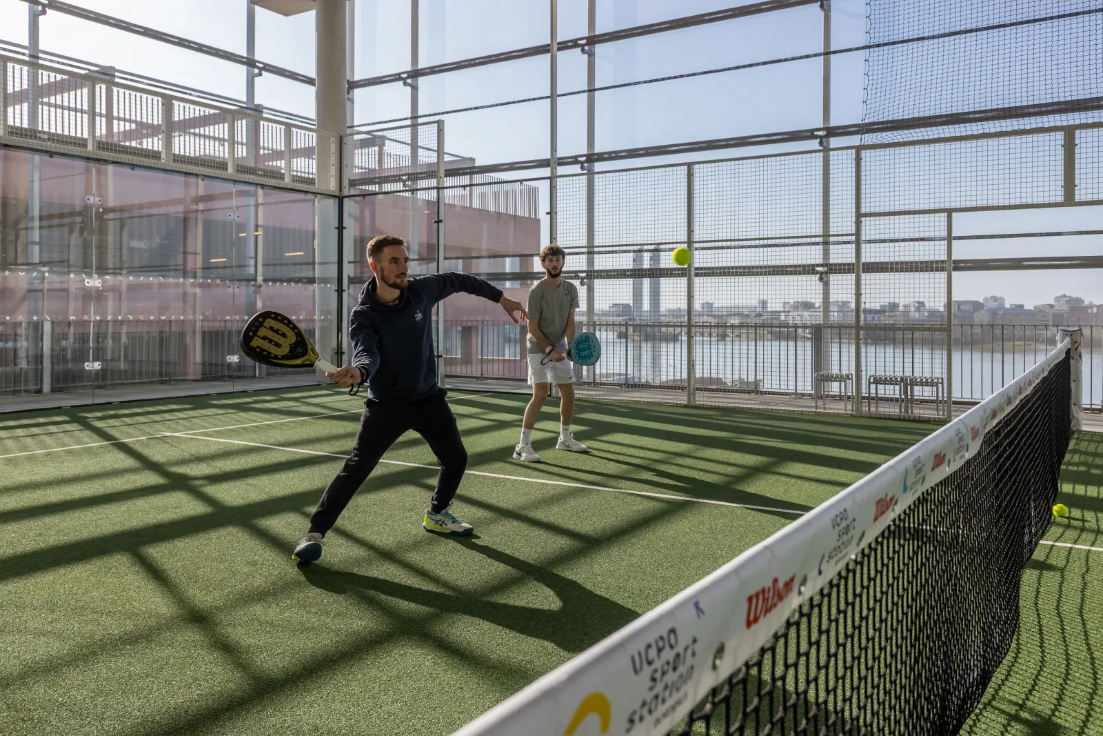 Two padel players on an elevated court in Bordeaux, one in playing position with his racket, the other waiting, with the city and Garonne river visible in the background through protective glass panels.