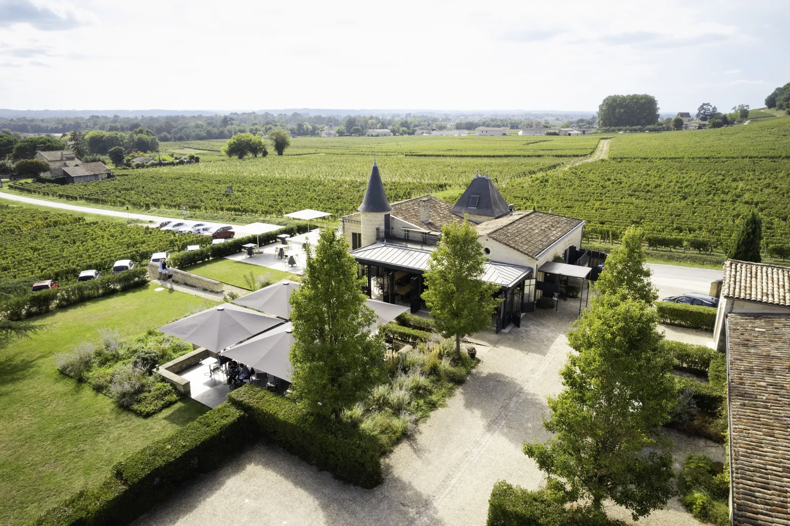 Aerial view of Château de Candale near Bordeaux, wine estate featuring conical-roofed buildings surrounded by vineyards, shaded terrace area and parking, rolling countryside with expansive vineyard landscape.