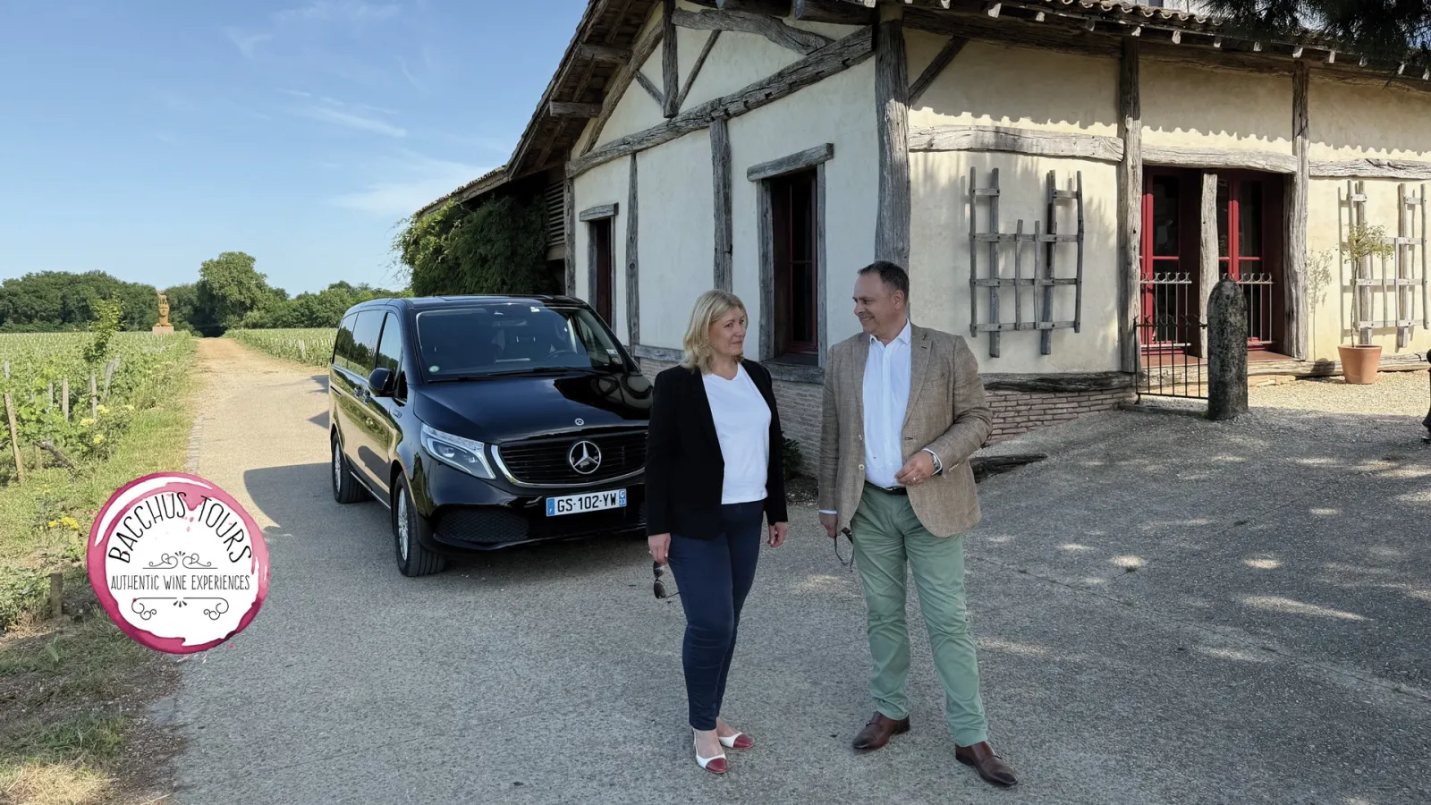 Two people standing in front of a traditional Bordeaux house with a black Mercedes vehicle, vineyards in the background, Bacchus Tours logo visible