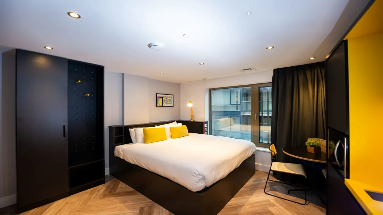Modern aparthotel bedroom featuring a queen-size bed, black furniture, yellow accents, large windows overlooking Bordeaux, and contemporary amenities.