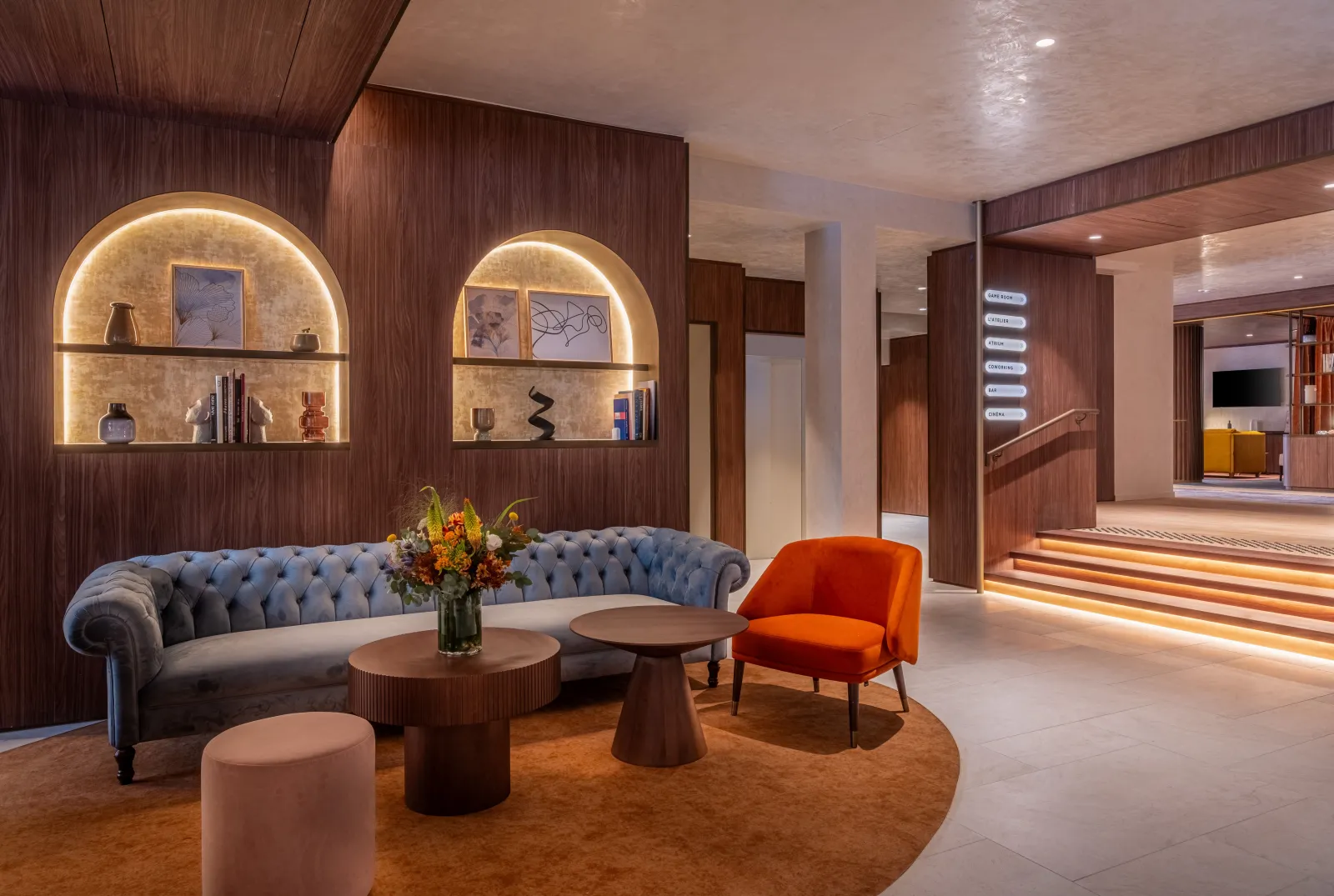 Elegant lobby of Hotel Burdigala in Bordeaux featuring dark wood paneling, gray Chesterfield sofa, orange accent chair, brown side tables, illuminated arched niches and terracotta-colored carpet