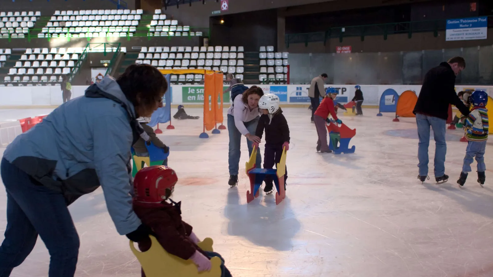 Children's ice skating lesson at Mériadeck Ice Rink in Bordeaux, with instructors assisting young skaters wearing helmets and using colorful learning aids on the ice.