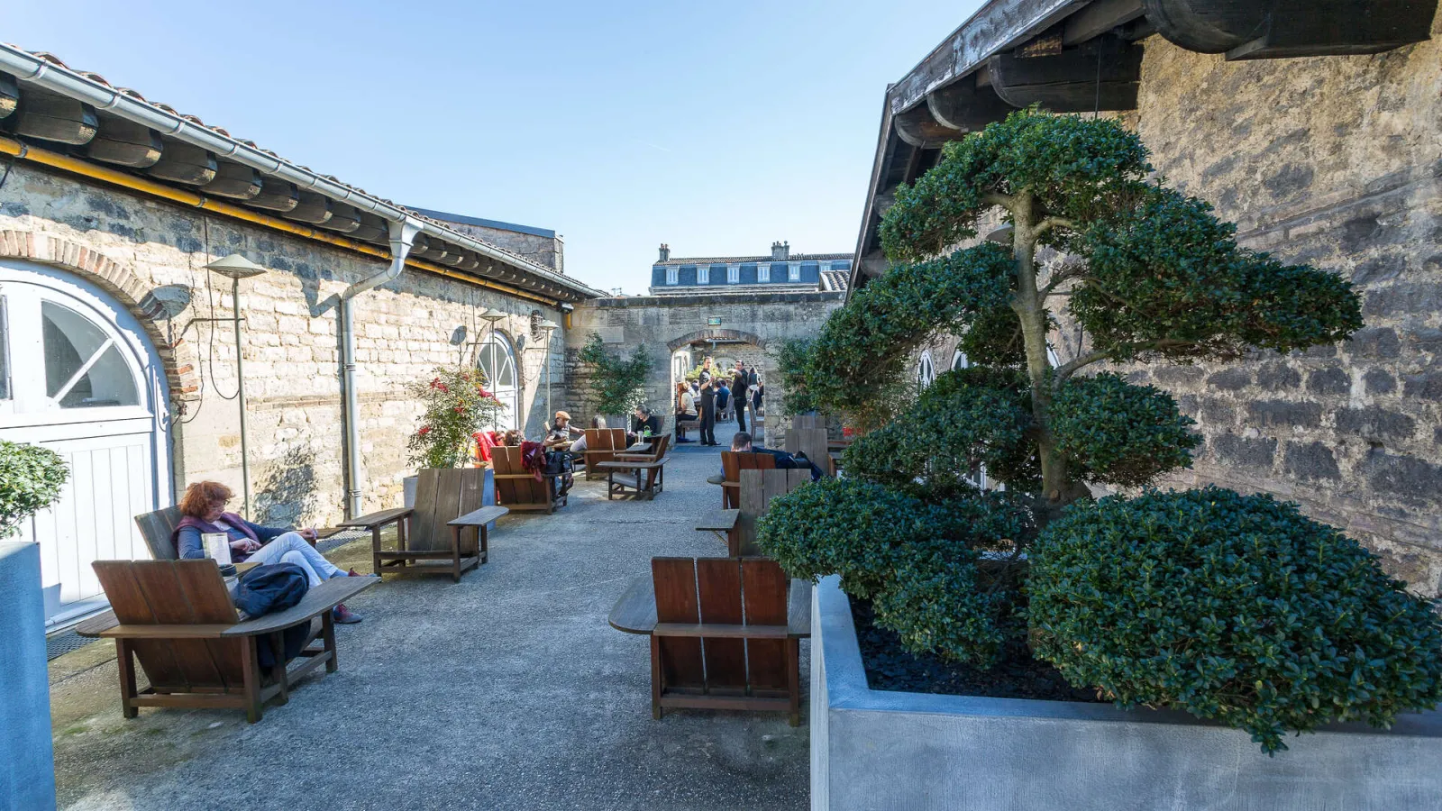 Interior courtyard of the CAPC Museum of Contemporary Art in Bordeaux featuring wooden lounge chairs, stone walls, ivy-covered facades, and visitors relaxing in a historic architectural space.
