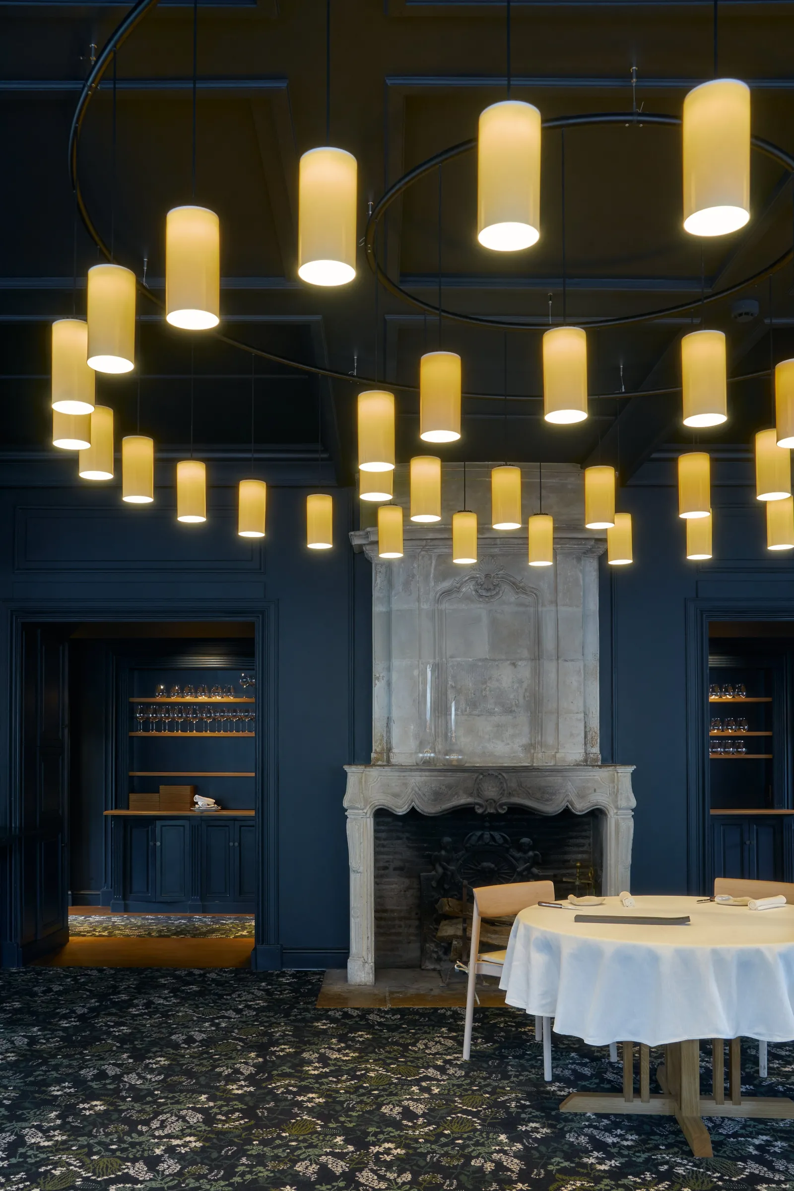 Elegant dining room at La Grand'Vigne restaurant in Bordeaux featuring golden cylindrical pendant lights on ceiling, dark blue walls, white marble fireplace and formally set round tables