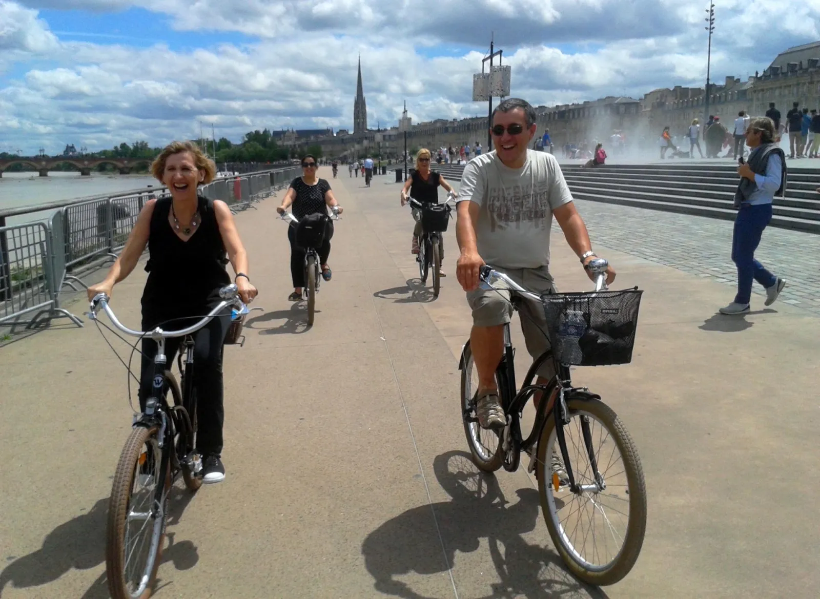 Group of cyclists riding on a riverside promenade in Bordeaux, with historic buildings and a fountain in the background