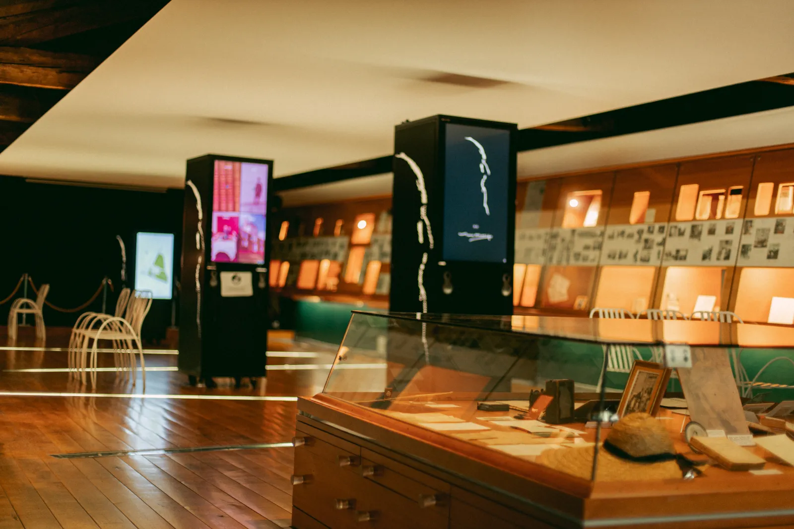 Interior of the Malagar Estate Museum in Bordeaux, featuring an exhibition with wooden display cases, interactive digital screens, white designer furniture, and an illuminated showcase of historical objects.