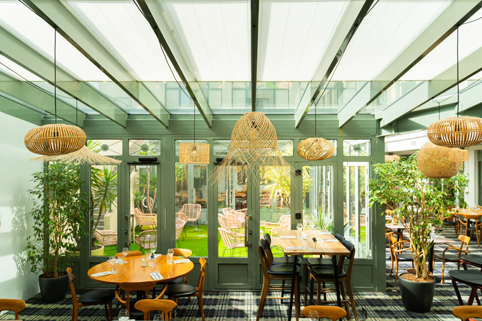 Modern restaurant dining room with glass conservatory, green structural beams, round wooden tables, colorful chairs and woven rattan pendant lights. Direct access to outdoor garden seating area.
