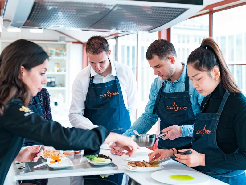 Four people wearing blue Côté Cours aprons participate in a cooking class in a modern kitchen with large windows overlooking Bordeaux. They are preparing dishes together at a work counter.