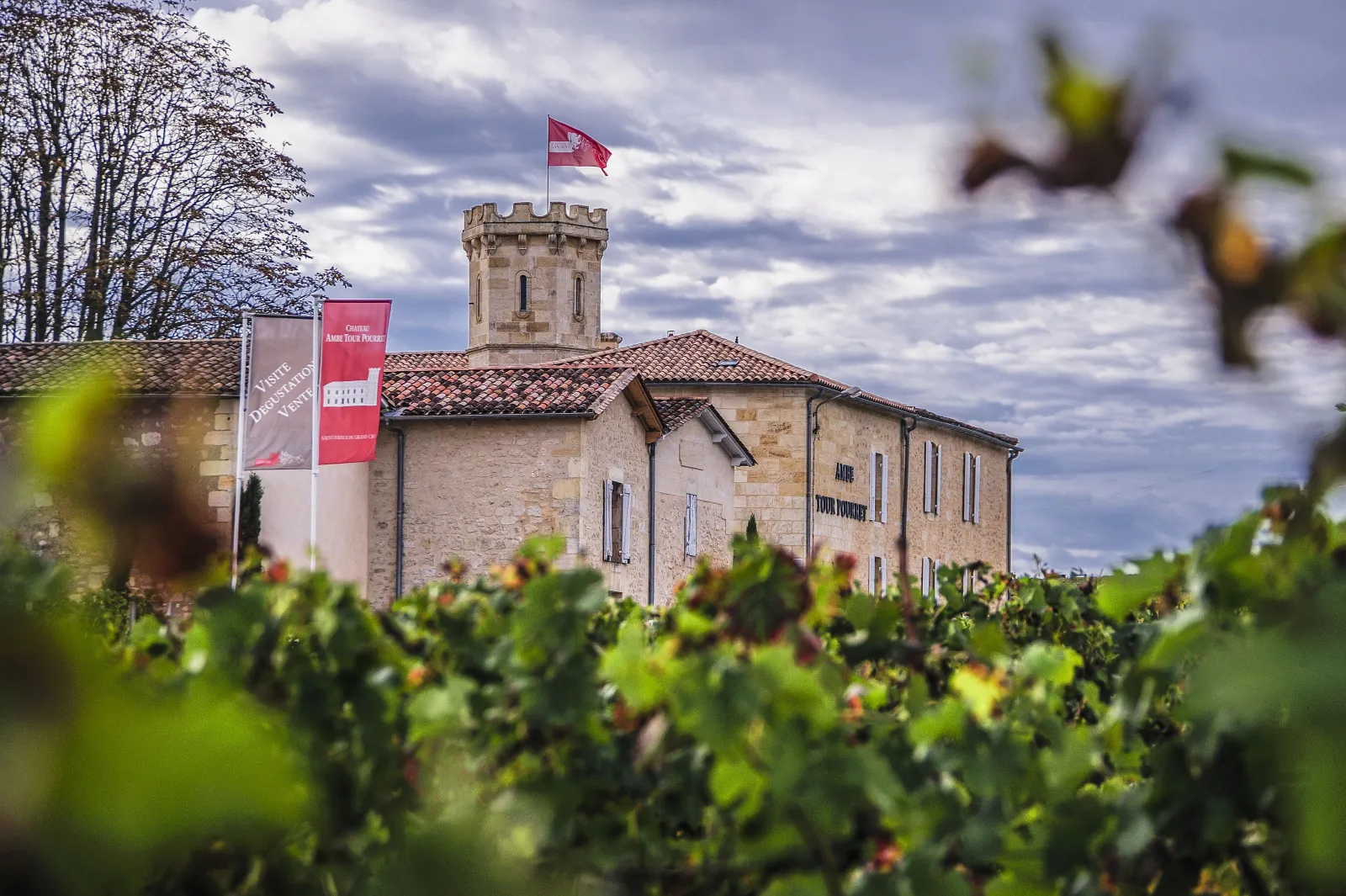 Ambe Tour Pourret Castle in Bordeaux, stone building with crenellated square tower, red and white flag, surrounded by vineyards in the foreground.