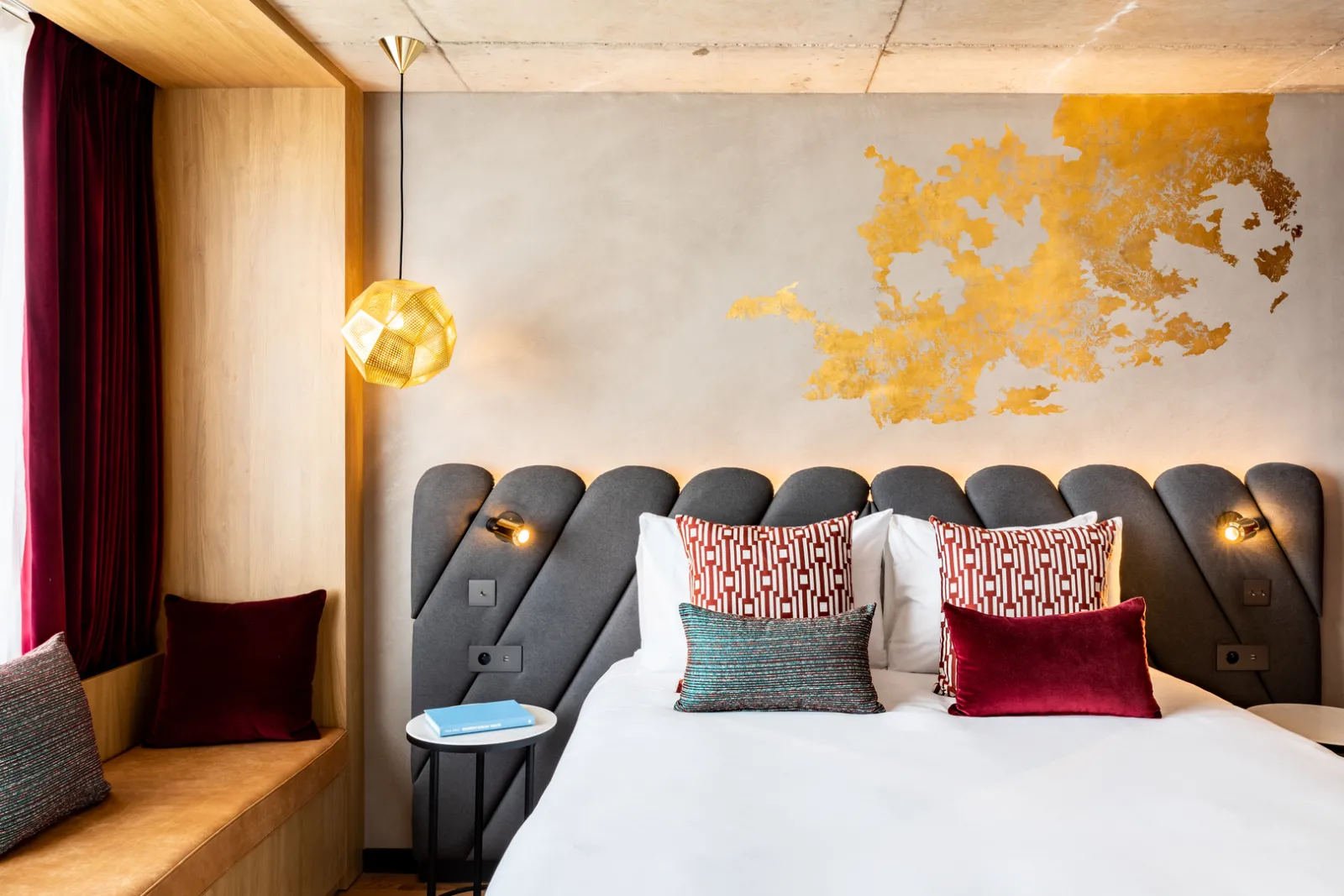 Modern hotel room at Renaissance Bordeaux featuring a king-size bed with gray tufted headboard, red and white decorative pillows, golden pendant lamp, a gold Bordeaux map on the wall, and wooden welcome nook.
