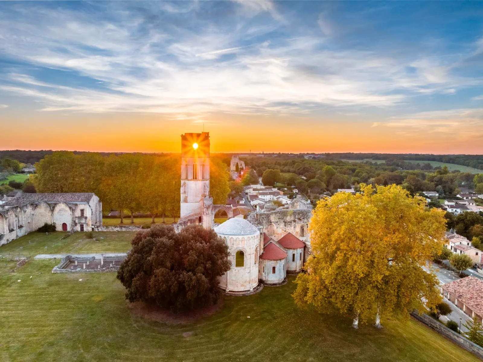 Aerial view of La Sauve-Majeure Abbey at sunset, featuring its illuminated bell tower, Romanesque buildings, and surrounding golden trees in the evening light