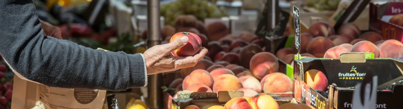 Person holding a peach at the Saint-Seurin farmers market in Bordeaux, with 'Fruites'font' fruit boxes and other peaches displayed in the background