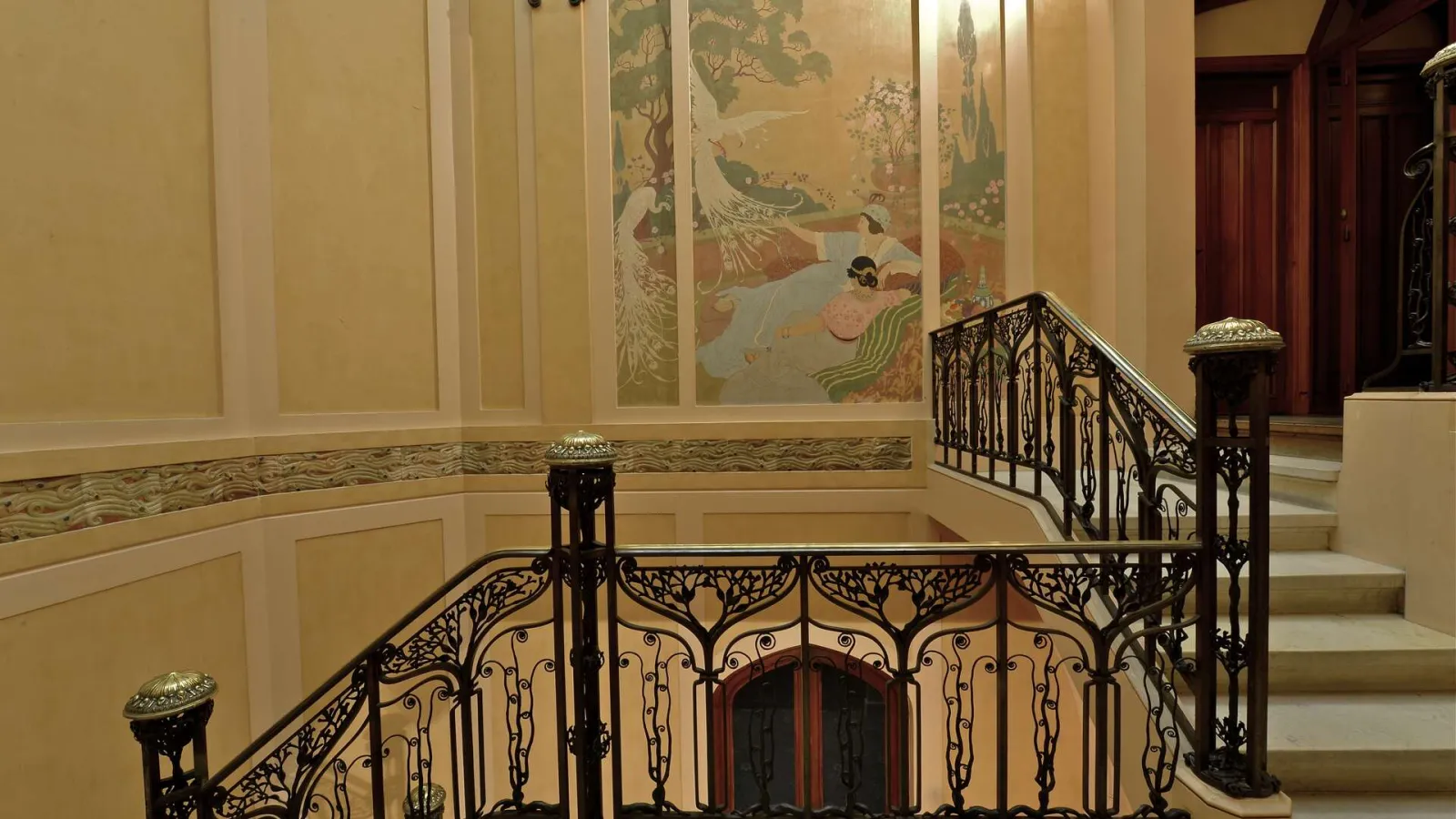Elegant staircase of the Frugès Hotel in Bordeaux featuring ornate wrought iron railing, beige paneled walls with golden friezes, pastoral mural artwork, and red doors