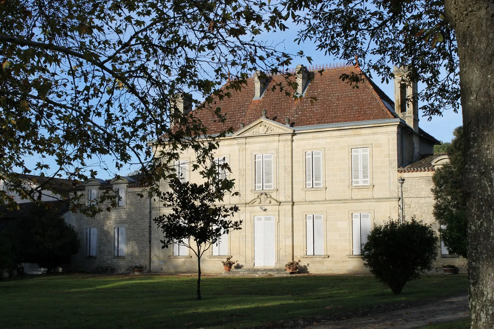 Château Mangot in Bordeaux, an elegant cream-colored stone mansion with red tile roof, surrounded by lawn and trees, featuring a symmetrical facade with white windows and central portico.