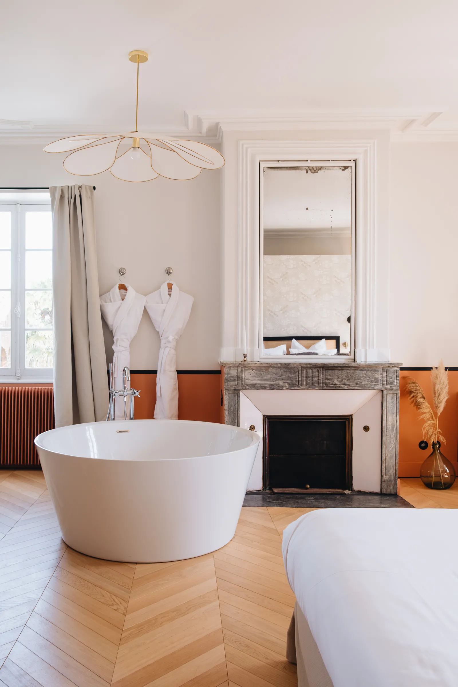 Luxurious bathroom at Château Réal in Bordeaux featuring a white freestanding bathtub, stone fireplace, framed mirror, geometric gold chandelier, and orange decorative accents.