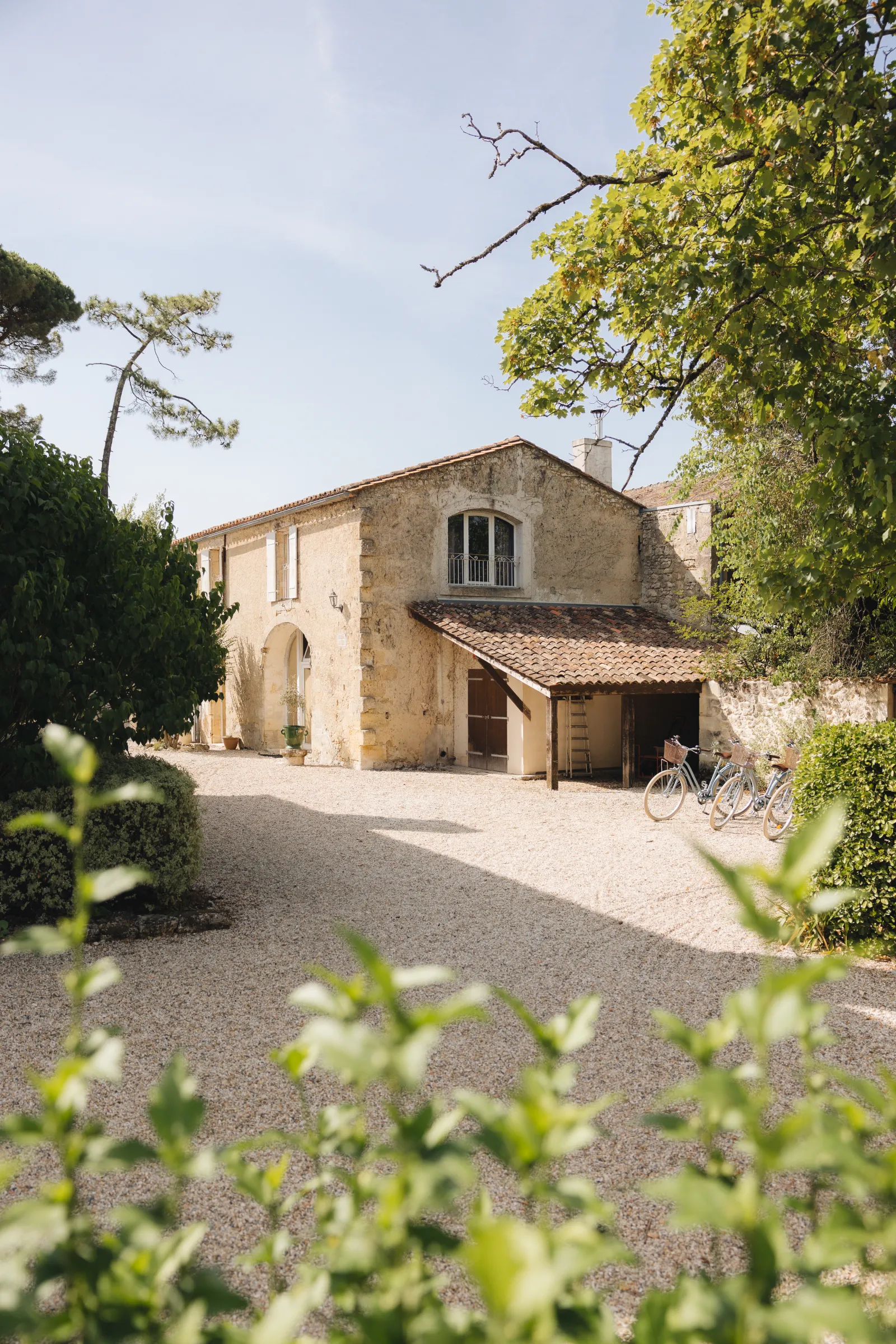 Stone facade of a Bordeaux château with two stories, wooden door, arched windows, surrounded by lush green vegetation and gravel driveway, with bicycles parked under the shelter.