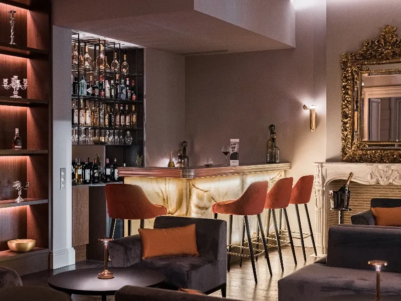 Elegant hotel bar featuring an illuminated counter, terracotta bar stools, bottle-lined shelves, ornate gold mirror, and modern furnishings in warm tones