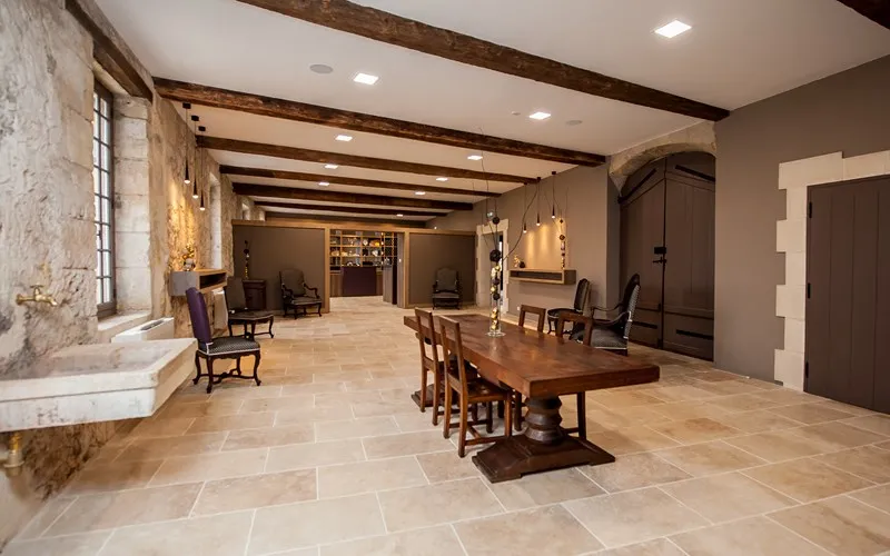 Vaulted reception hall of Château de Portets in Bordeaux featuring exposed wooden beams, wooden dining table, antique chairs, stone flooring, warm lighting and access to adjoining rooms