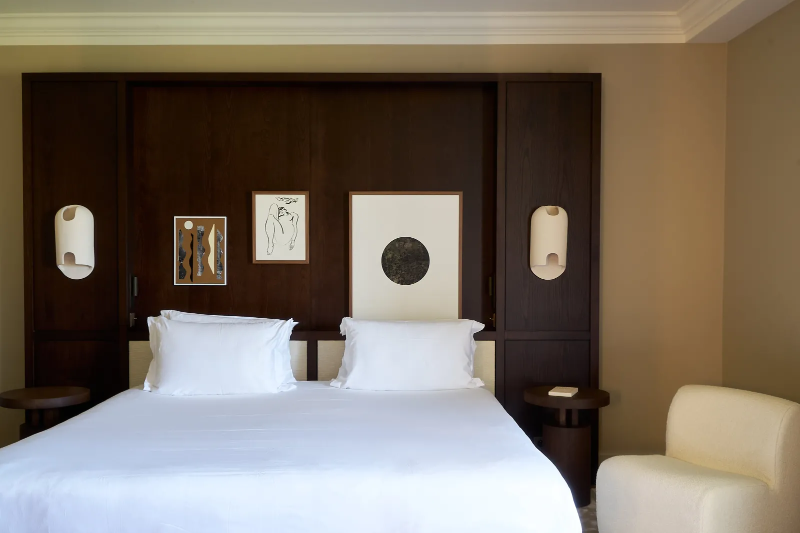 Elegant hotel bedroom at Les Sources de Caudalie in Bordeaux, featuring a white bed, dark wood headboard with modern artworks, wall sconces, and a cream armchair.