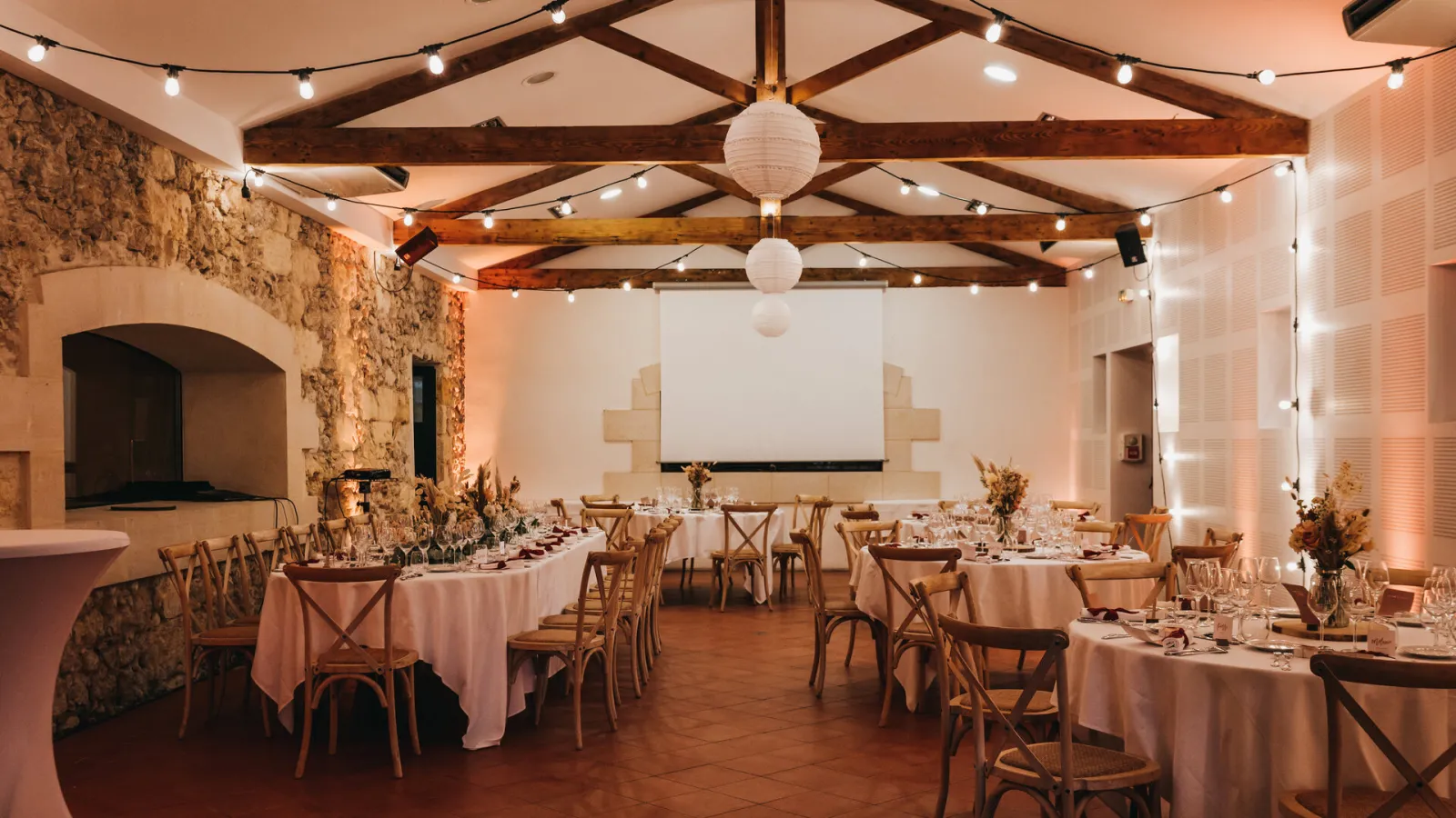 Elegant reception hall at Château Saint Ahon in Bordeaux featuring round tables with white linens, wooden chairs, exposed beams, warm ambient lighting and projection screen