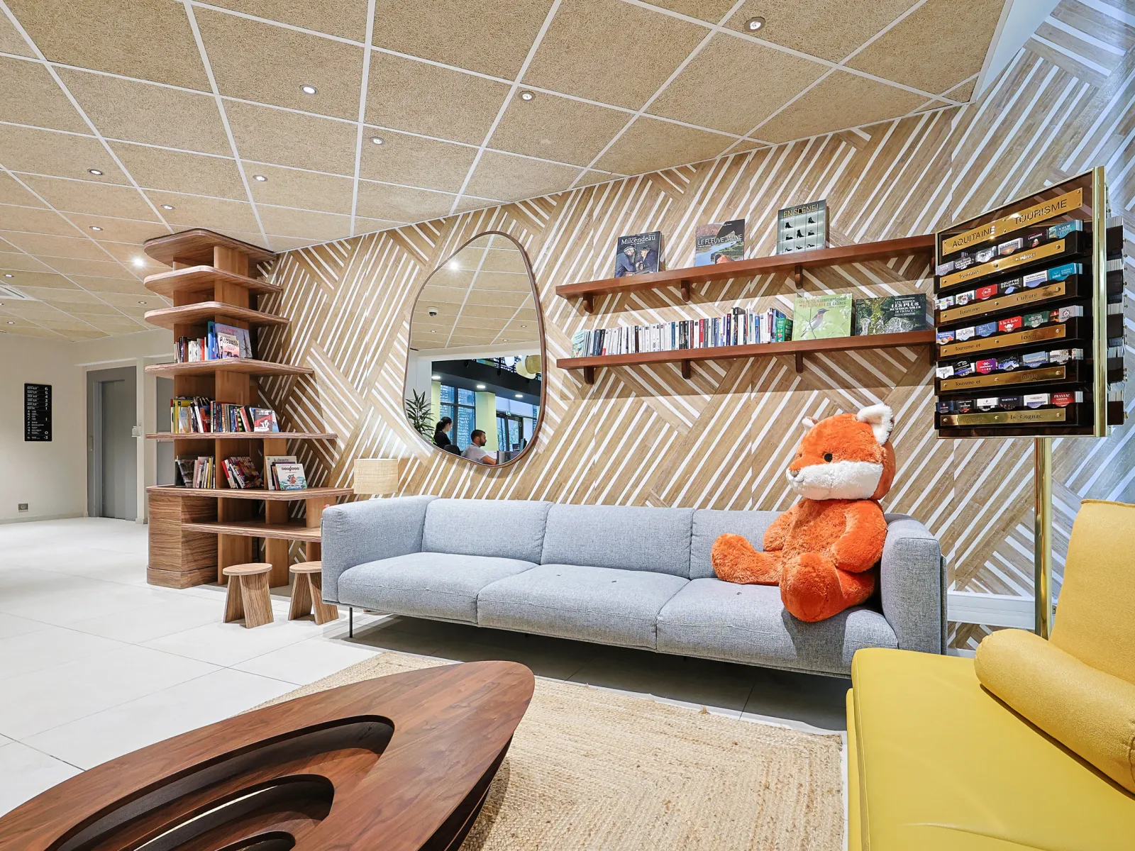 Modern living area of a serviced apartment featuring grey sofa, wooden shelves, round mirror, orange teddy bear and contemporary furniture in Bordeaux