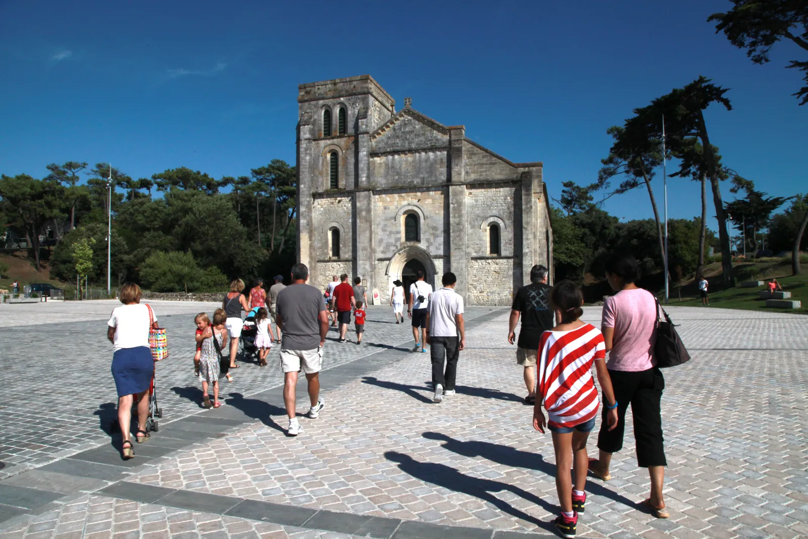 Visitors strolling on the paved square in front of Bordeaux's Saint-Michel Basilica, a Gothic stone religious building with a square tower, surrounded by trees under blue sky.
