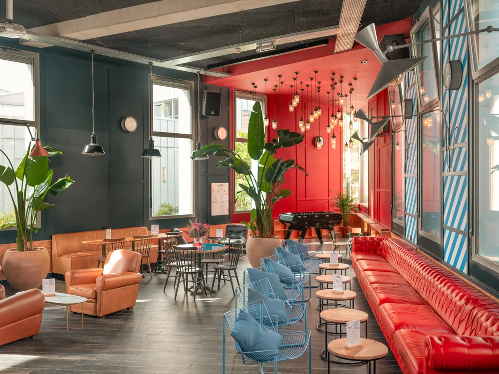 Modern living space at Eklo Bordeaux featuring a vibrant lounge with red and teal walls, orange armchairs, red sofa, blue chairs, green plants, pendant lights and large windows.