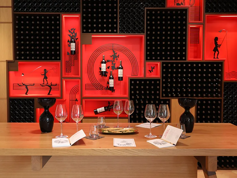 Wine tasting at Château Marquis d'Alesme Becker in Bordeaux: wooden table with glasses and bottles in front of a modern wall featuring black and red panels with geometric patterns and silhouettes.