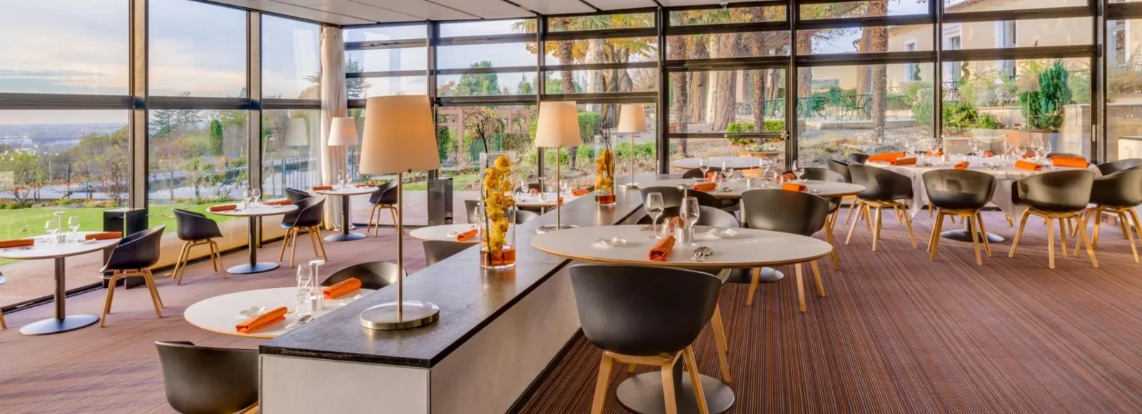 Panoramic dining room at Saint-James restaurant in Bordeaux featuring floor-to-ceiling windows overlooking the valley, modern round and rectangular tables with black chairs with wooden legs, orange accents, and outdoor landscaped terrace.