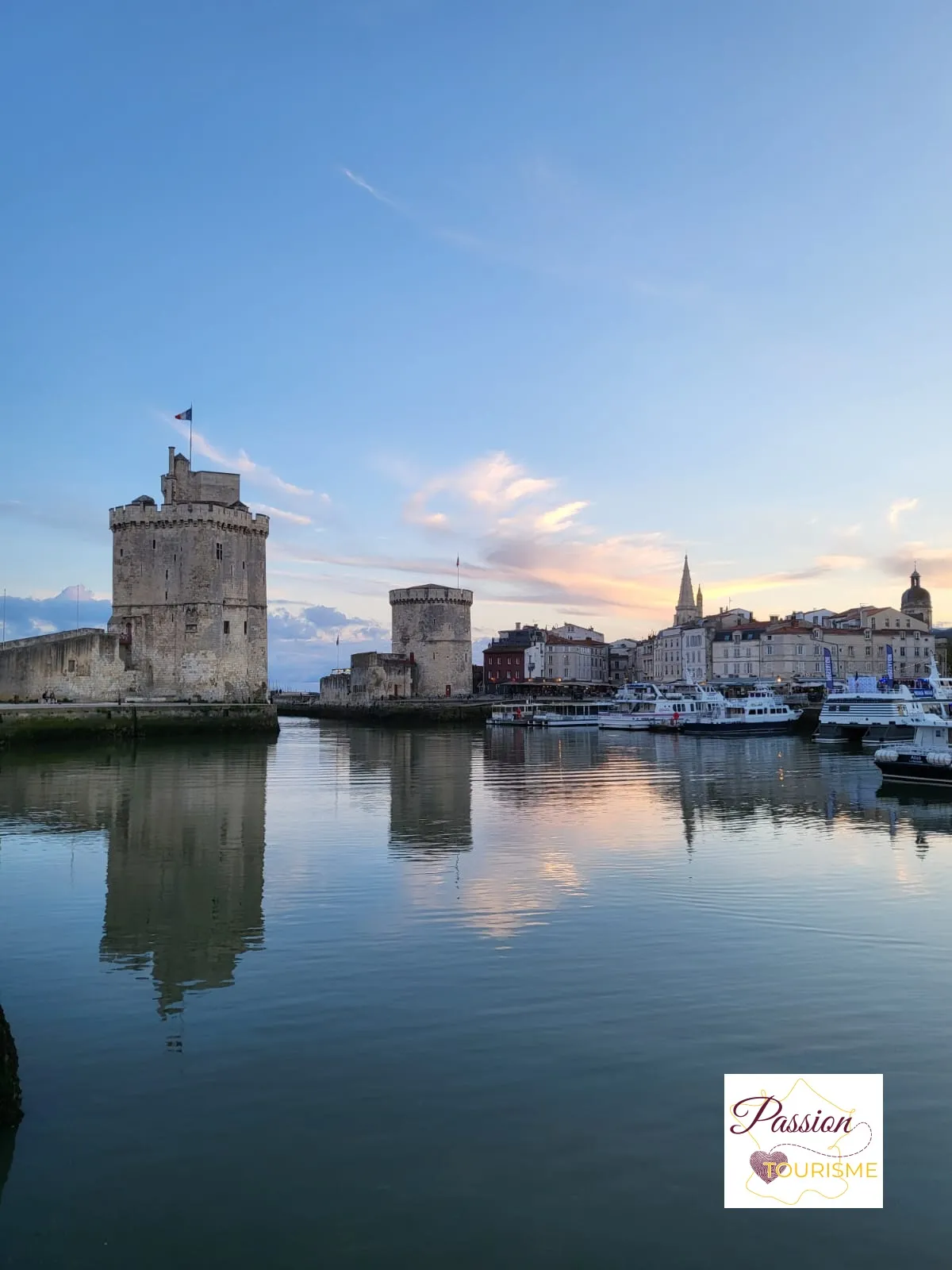 Panoramic view of La Rochelle's harbor featuring three historic towers reflected in calm water, the old town in the background and a colorful sunset