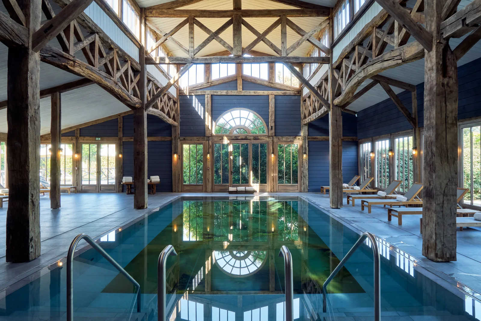 Indoor swimming pool at Les Sources de Caudalie in Bordeaux, featuring exposed wooden beams, large windows overlooking greenery, and substantial columns