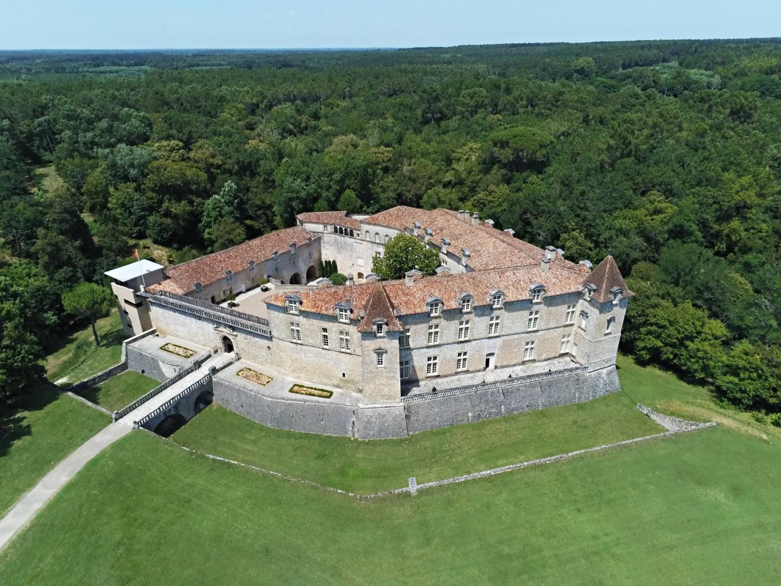 Aerial view of Château Royal de Cazeneuve, a medieval fortress surrounded by moats and ramparts with red tile roofs, nestled in lush green forest landscape near Bordeaux.