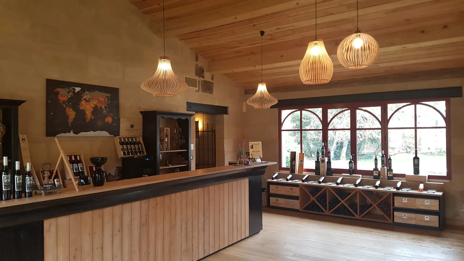 Elegant wine tasting room interior with wooden counter, wine bottle displays, light wooden ceiling, modern pendant lights, and large windows overlooking the vineyard grounds.