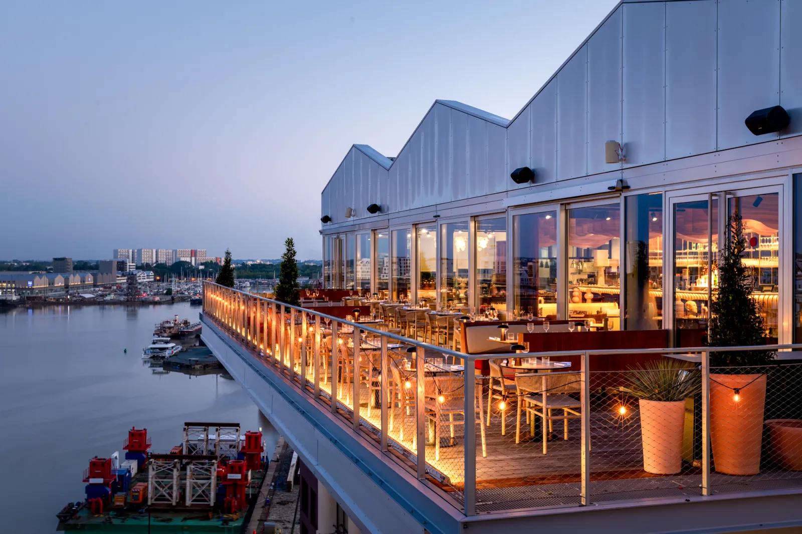 Modern riverside restaurant in Bordeaux with an illuminated terrace overlooking the Garonne at dusk, featuring contemporary architecture with sawtooth roof and large glass windows.