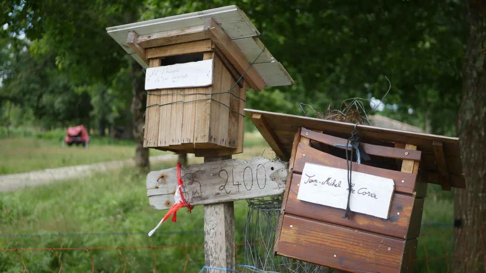 Two handmade wooden birdhouses mounted on a post at the Bergerie des Matruques in Bordeaux, with handwritten labels and a green landscape in the background.