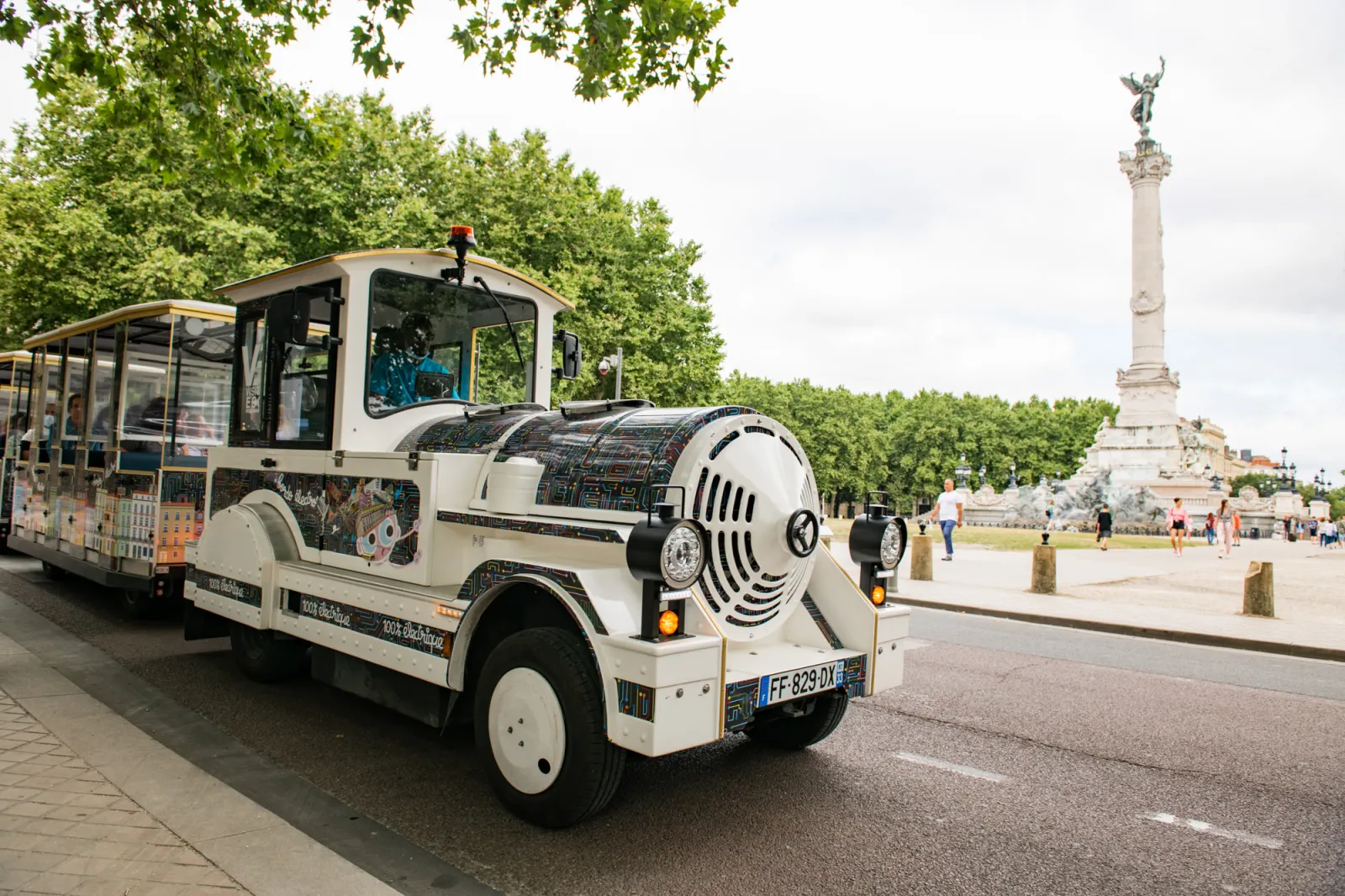 Bordeaux's electric tourist train, a white miniature train with a stylized steam locomotive design, running in front of the Monument to the Girondins in Bordeaux's Place de la Bourse.
