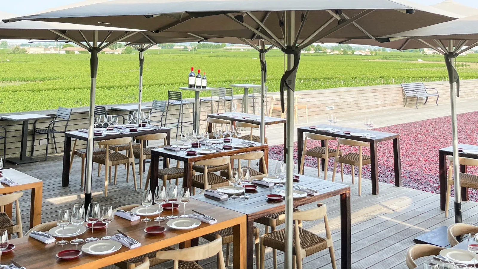 Outdoor restaurant terrace with set tables, grey umbrellas, vineyards in the background and red floral decoration