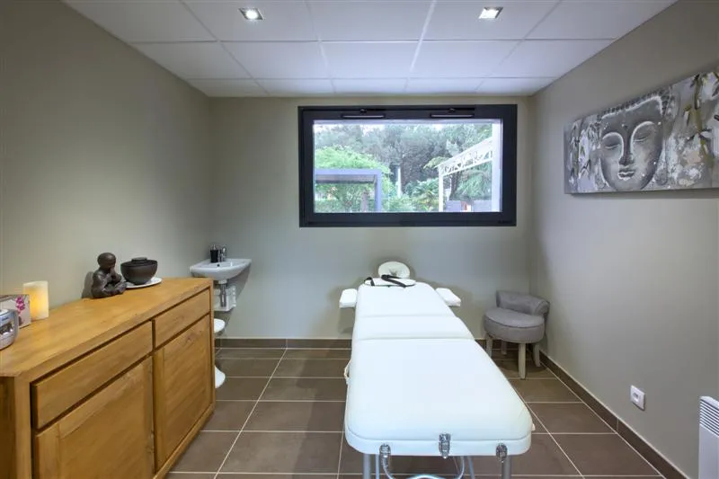 Wellness and treatment room featuring a white massage table, wooden furniture, sink, zen decoration, and window overlooking the garden at Au Comté d'Ornon hotel in Bordeaux.