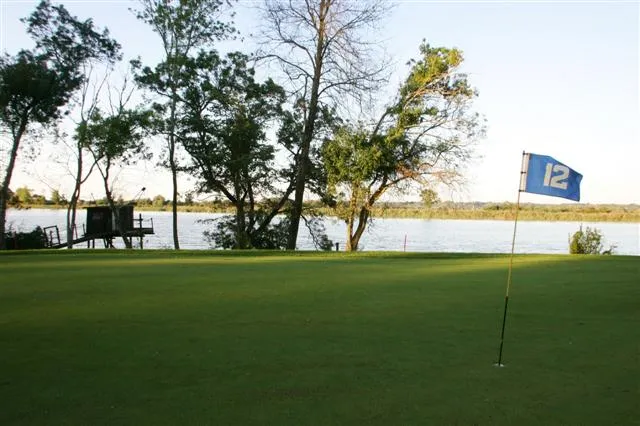 Golf course with flag at hole 12, overlooking lake and trees, hotel terrace in background